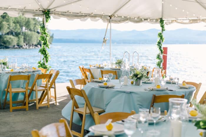 North Dock set for a dinner with light blue tablecloths and lemon place settings, beyond the tent a beautiful view of Lake Champlain and the Adirondacks.