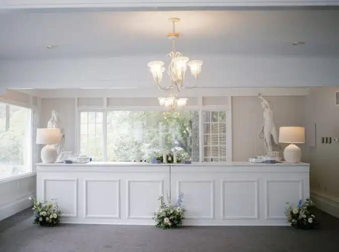 A white wooden bar decorated with flower arrangements placed in the front. Next to the bar, on either side, are stone statues.